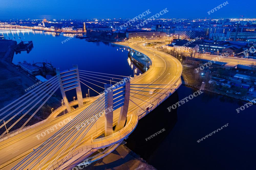 На фотографии изображён мост Бетанкура, один из новейших мостов Санкт-Петербурга. Мост соединяет Васильевский остров с Приморским районом и отличается своей современной конструкцией с вантовой поддержкой. Вечером мост освещён, и его огни отражаются в спокойных водах реки, создавая красивую картину города.
